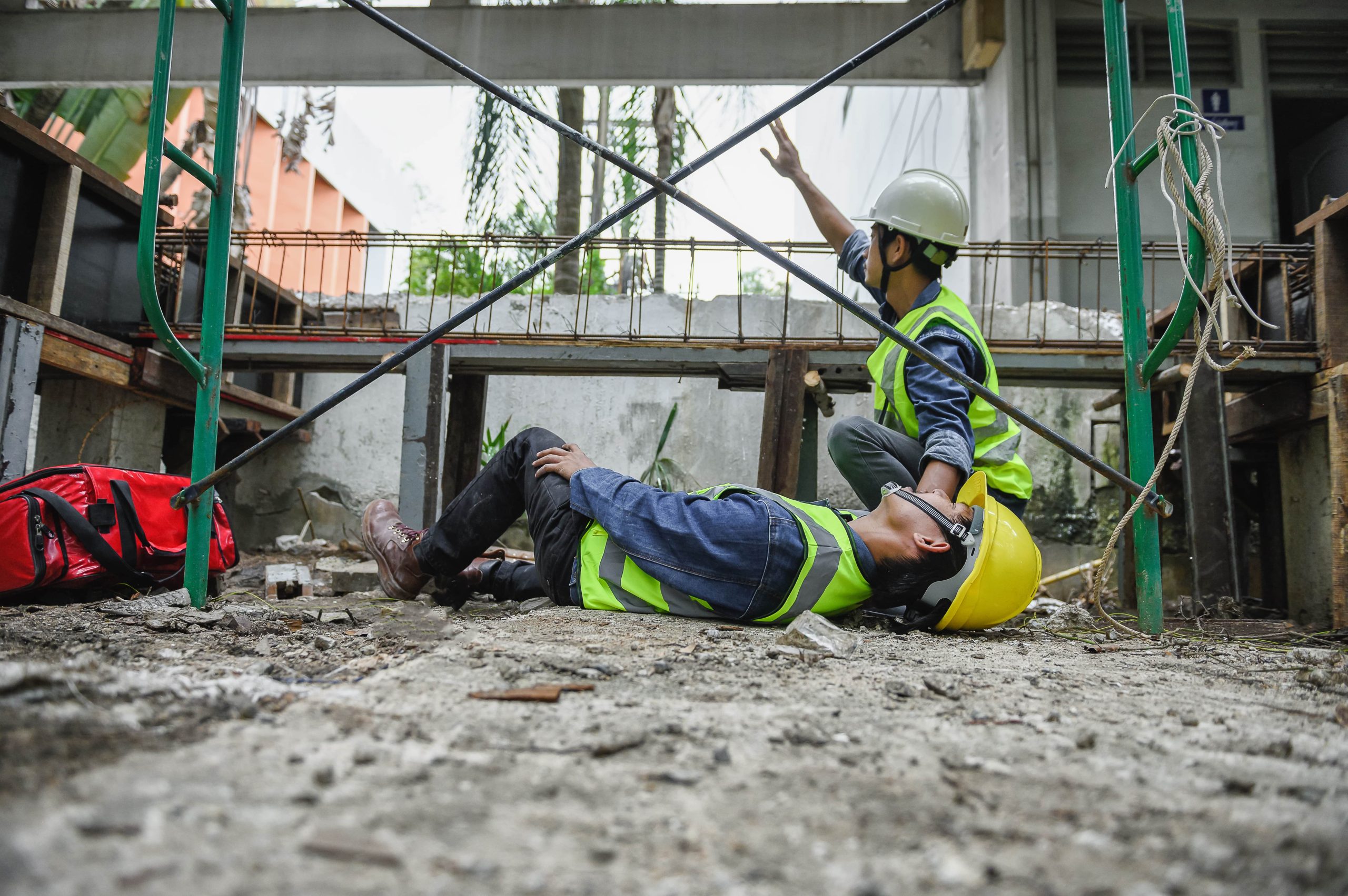 Due operai edili con giubbetti ad alta visibilità e caschi di sicurezza in un cantiere; uno di loro è a terra privo di sensi, mentre l'altro chiede aiuto con urgenza.